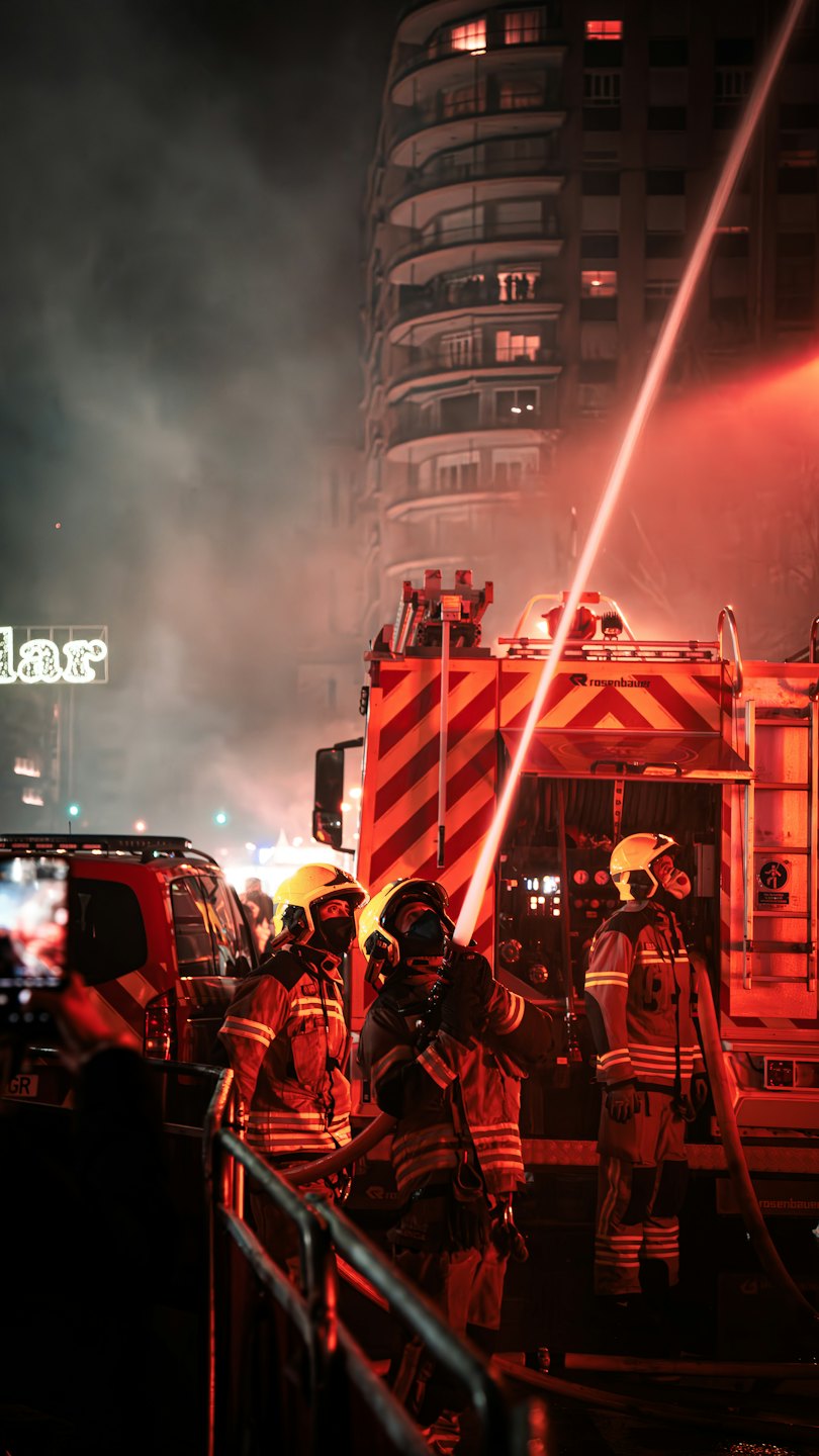 Drie brandweermannen spuiten water uit een slang op een gebouw.